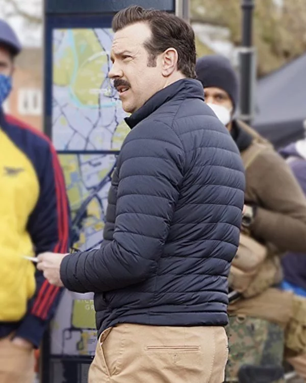 Side profile view of Jason Sudeikis wearing the dark navy blue Ted Lasso puffer jacket with horizontal quilting and stand-up collar
