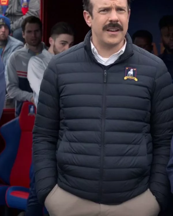 Front view of Jason Sudeikis as Ted Lasso wearing the dark navy blue puffer jacket featuring horizontal quilting, stand collar, and AFC Richmond chest crest. Jason Sudeikis Ted Lasso Puffer Jacket